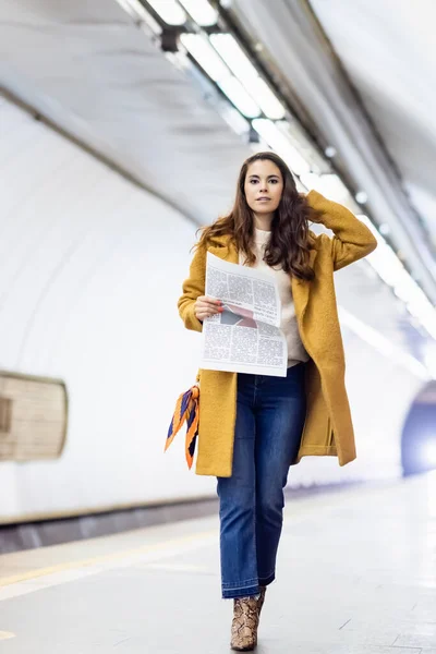 Giovane donna in elegante vestito autunno guardando la fotocamera mentre tiene il giornale sulla stazione della metropolitana — Foto stock