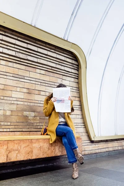 Frau im schicken Herbst-Outfit verdeckt Gesicht mit Zeitung auf U-Bahn-Bahnsteigbank — Stockfoto