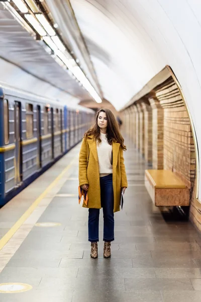 Donna elegante in abiti autunnali guardando treno su piattaforma sotterranea, sfondo sfocato — Foto stock
