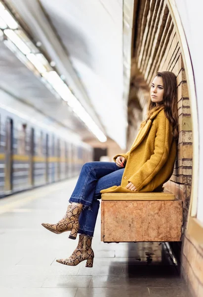 Вдумлива жінка в осінньому вбранні, сидячи на лавці платформи метро з розмитим поїздом — стокове фото