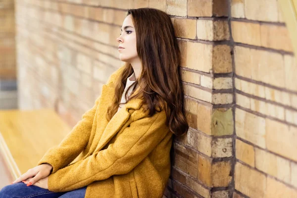 Вдумчивая женщина в осеннем пальто сидит на скамейке в метро на размытом фоне — стоковое фото