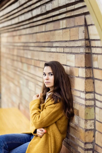Young woman in autumn coat looking at camera while sitting on subway platform bench on blurred background — Stock Photo
