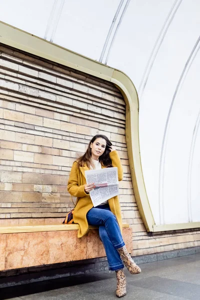 Young woman in stylish autumn clothes touching hair while holding newspaper on metro platform bench - foto de stock
