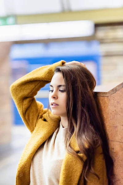 Молодая женщина в осеннем наряде трогает волосы, стоя у стены на станции метро — стоковое фото