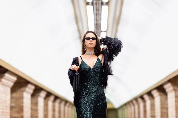 Trendy woman in black lurex dress and sunglasses posing at underground station with bottle of wine — Stock Photo