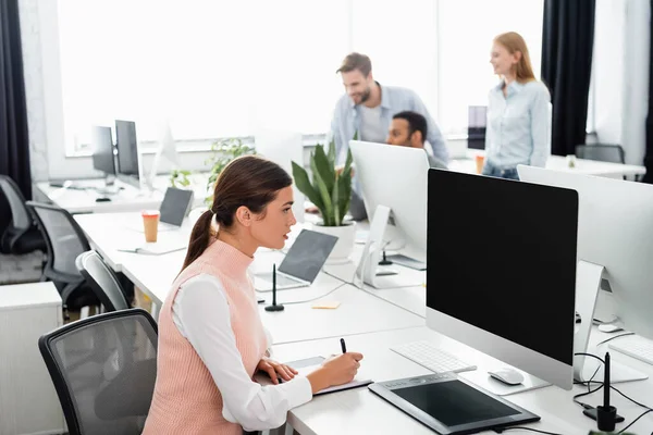 Empresaria escribiendo en un cuaderno cerca de la computadora, tableta gráfica y colegas multiétnicos en un fondo borroso en la oficina — Stock Photo