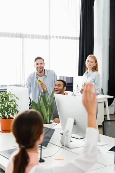 Smiling multiethnic businesspeople looking at colleague with raised hand on blurred foreground in office — Stock Photo