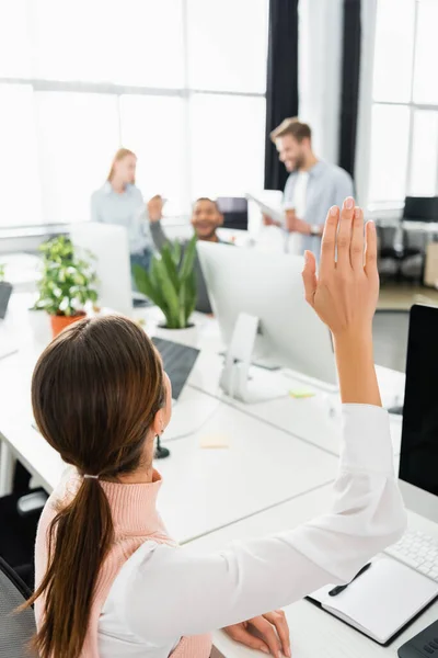 Geschäftsfrau mit erhobener Hand sitzt neben Computern und Kollegen auf verschwommenem Hintergrund im Büro — Stockfoto