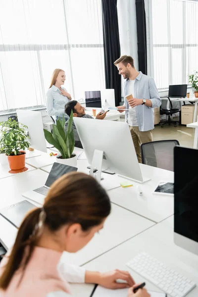Colegas multiétnicas hablando cerca de dispositivos y empresaria escribiendo en cuaderno en primer plano borroso - foto de stock