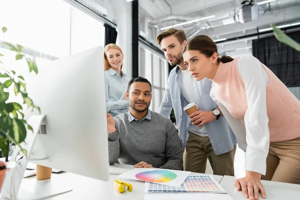 Empresários multiétnicos olhando para monitor de computador em primeiro plano borrado perto de amostras coloridas na mesa — Fotografia de Stock