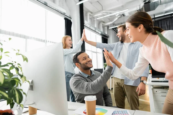 Alegre gente de negocios multicultural dando alta cinco cerca de la computadora y muestras de colores en primer plano borrosa en la oficina - foto de stock