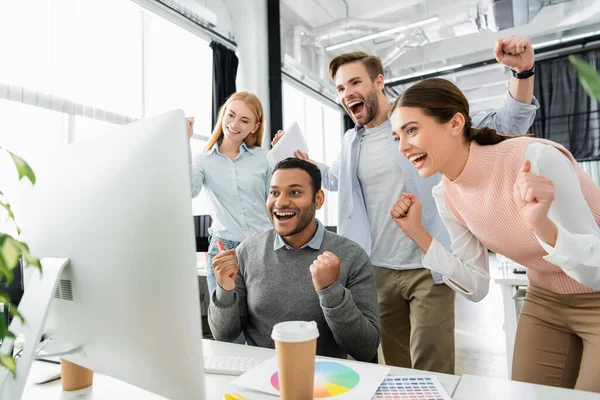 Cheerful multiethnic businesspeople showing yeah gesture near computer, colorful swatches and coffee to go on blurred foreground — Stock Photo