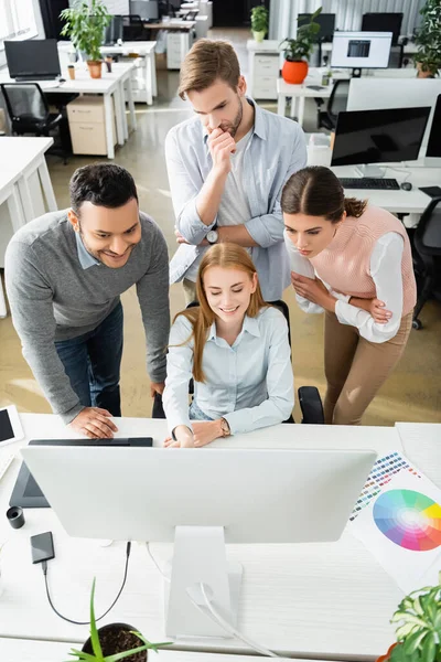 Vista de ángulo alto de los empresarios multiétnicos sonrientes y pensativos que utilizan la computadora cerca de muestras coloridas en primer plano borroso - foto de stock