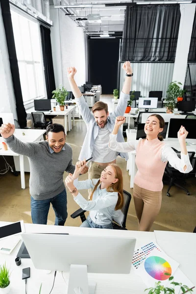 Fröhliche multikulturelle Geschäftsleute mit Ja-Geste in der Nähe von Computern und bunten Mustern auf dem Bürotisch — Stockfoto