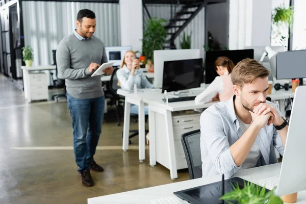 Uomo d'affari che utilizza computer vicino tablet grafica e colleghi multietnici su sfondo sfocato in ufficio — Foto stock