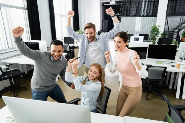 Positive multiethnic businesspeople showing yeah gesture near computer on blurred foreground in office — Stock Photo