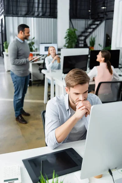 Pensive businessman looking at computer near graphics tablet and multicultural colleagues on blurred background — Stock Photo