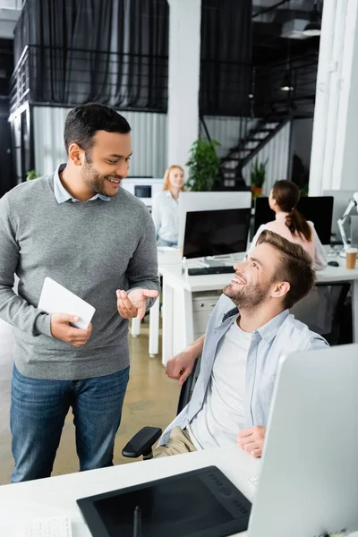 Empresario indio con tableta digital hablando con colega cerca de gráficos tableta y computadora en primer plano borroso en la oficina - foto de stock