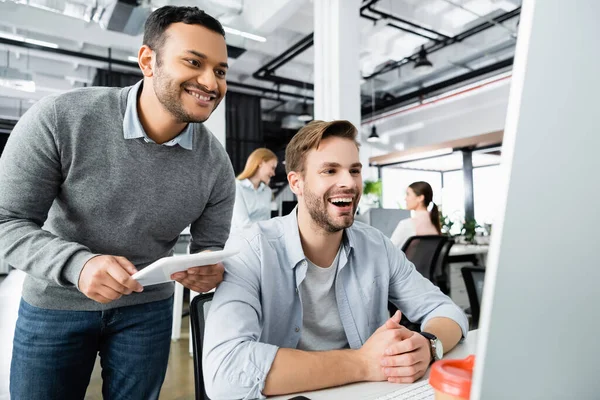 Cheerful multiethnic businessmen with digital tablet looking at computer monitor on blurred foreground in office — Stock Photo