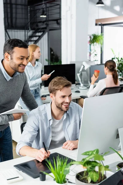 Sonrientes colegas multiétnicos utilizando gráficos tableta y computadora cerca de las plantas en primer plano borroso en la oficina - foto de stock