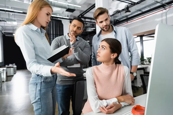 Geschäftsfrau mit Notizbuch zeigt auf Computer in der Nähe multiethnischer Kollegen im Büro — Stockfoto