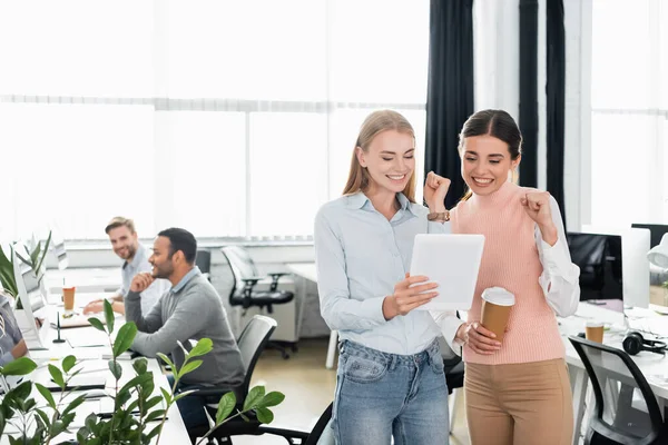 Smiling businesswomen showing yeah while holding digital tablet and coffee to go in office — Stock Photo