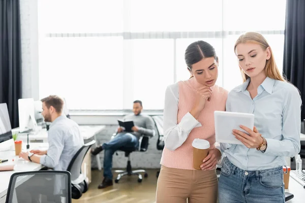 Businesswomen with coffee to go looking at digital tablet near colleagues in office — Stock Photo