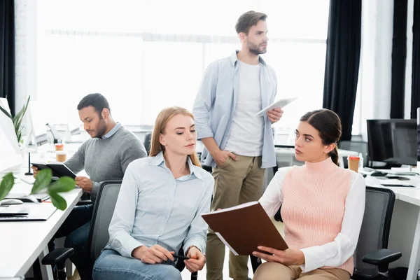 Geschäftsfrau hält Papiermappe in der Hand, während sie in der Nähe multiethnischer Kollegen im Büro im unscharfen Vordergrund arbeitet — Stockfoto