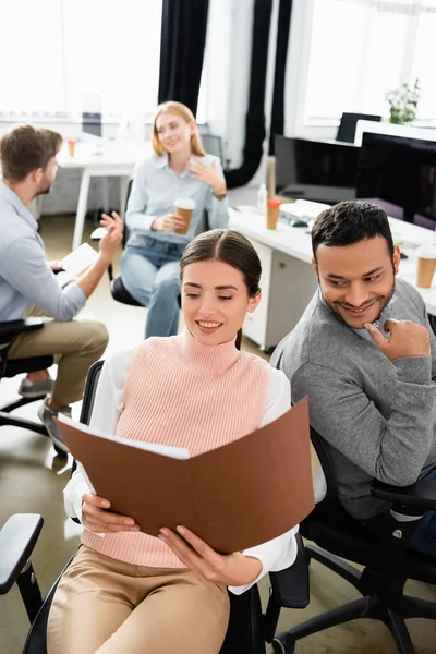 Negocios multiétnicos sonrientes mirando la carpeta de papel cerca de colegas con un fondo borroso en la oficina - foto de stock