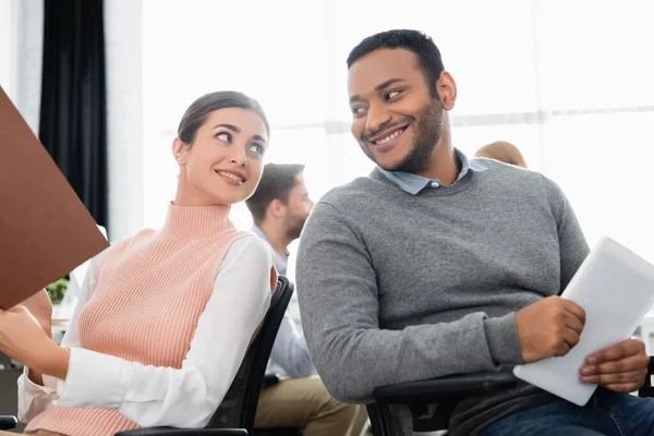 Mujer de negocios sonriente con carpeta de papel mirando a colega indio con tableta digital en la oficina - foto de stock