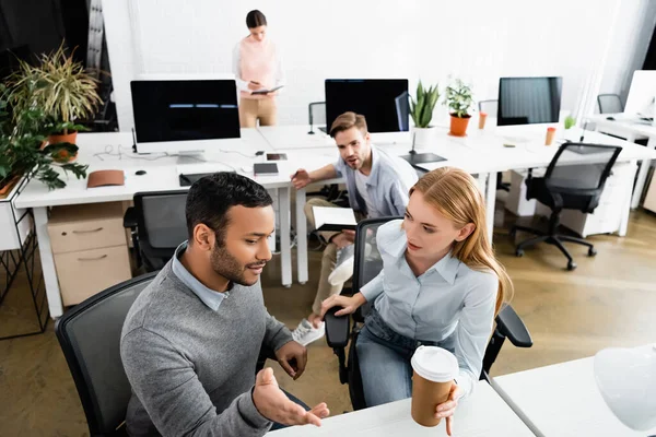 Empresario indio apuntando con la mano cerca de la empresaria con café para llevar y colegas sobre fondo borroso — Stock Photo