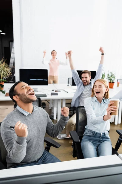 Emocionado empresario indio mostrando sí gesto cerca de colegas y computadoras en fondo borroso en la oficina - foto de stock