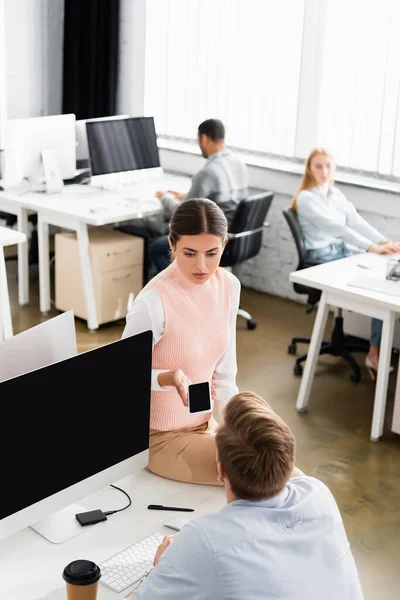 Femme d'affaires tenant smartphone avec écran blanc près collègue et ordinateur au bureau — Photo de stock