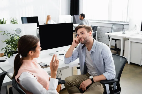 Businessman looking at colleague with coffee to go near devices in office — Stock Photo