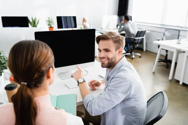 Empresário sorridente apontando com o dedo para monitor de computador perto de colega e café para ir no escritório — Fotografia de Stock