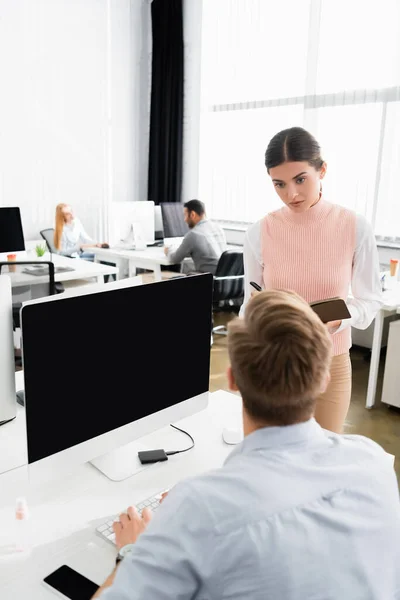 Femme d'affaires avec ordinateur portable debout près d'un collègue en utilisant l'ordinateur au premier plan flou dans le bureau — Photo de stock