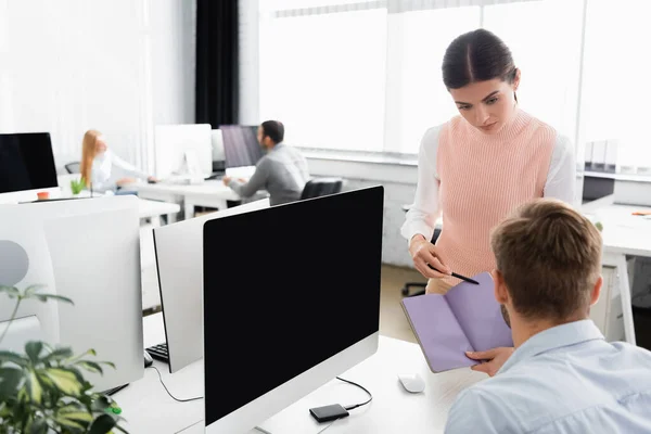 Femme d'affaires pointant du doigt un ordinateur portable près d'un collègue et d'un ordinateur avec un écran vide au premier plan flou — Photo de stock