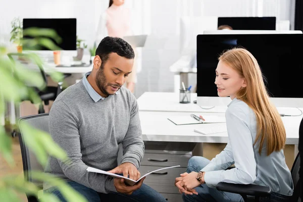 Indian businessman holding paper folder near smiling colleague and computer in office — Stock Photo