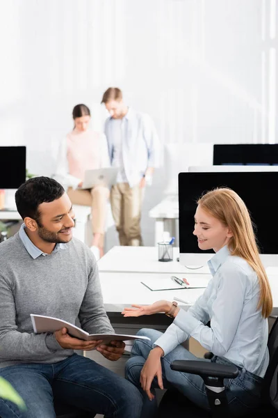 Empresários multiétnicos sorridentes olhando para a pasta de papel, enquanto colegas trabalhando em fundo turvo no escritório — Fotografia de Stock