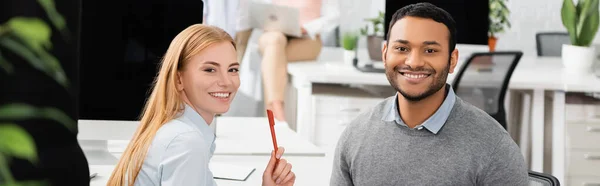 Donna d'affari sorridente con penna che guarda la macchina fotografica vicino al collega indiano in ufficio, striscione — Foto stock