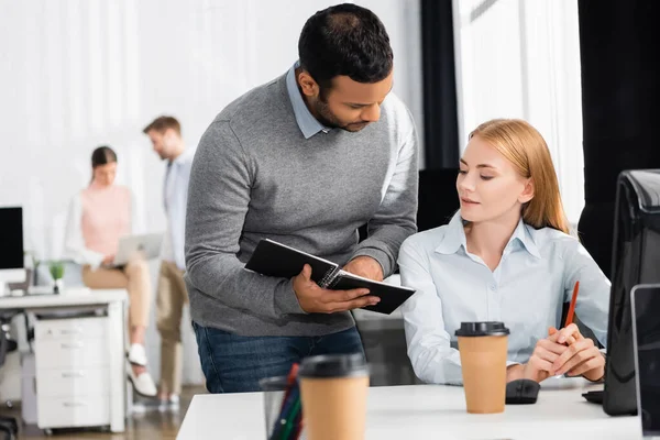 Indian businessman holding notebook near businesswoman and coffee to go on blurred foreground — Stock Photo