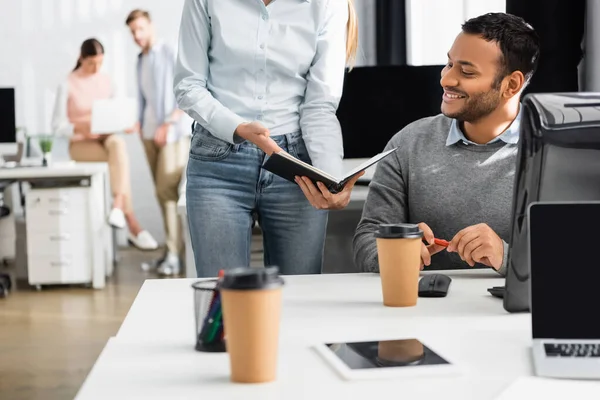 Smiling indian businessman sitting near businesswoman holding notebook and gadgets on blurred foreground in office — Stock Photo