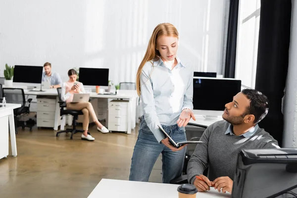 Businesswoman pointing at notebook near indian colleague and computer monitor on blurred foreground — Stock Photo