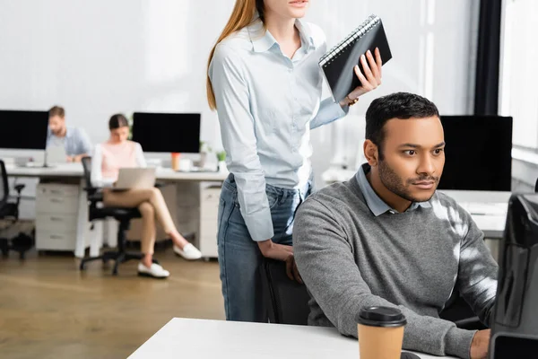 Hombre de negocios indio mirando el monitor de la computadora cerca de la mujer de negocios con cuaderno y café para ir sobre fondo borroso - foto de stock