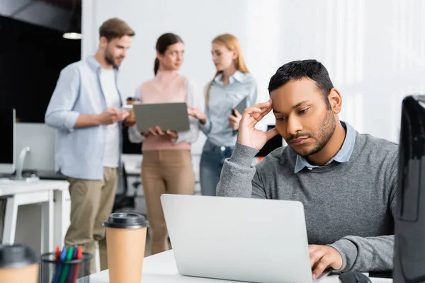 Concentrated indian businessman using laptop near coffee to go and colleagues on blurred background in office — Stock Photo