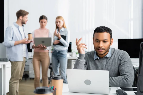 Indian businessman using laptop near coffee to go and colleagues working on blurred background in office — Stock Photo