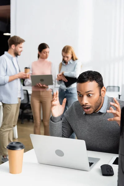 Schockierter indischer Geschäftsmann schaut auf Laptop, während Kollegen im Büro an verschwommenem Hintergrund arbeiten — Stockfoto