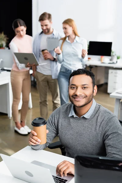 Улыбающийся индийский бизнесмен держит кофе, чтобы пойти, используя ноутбук рядом с коллегами на размытом фоне — стоковое фото