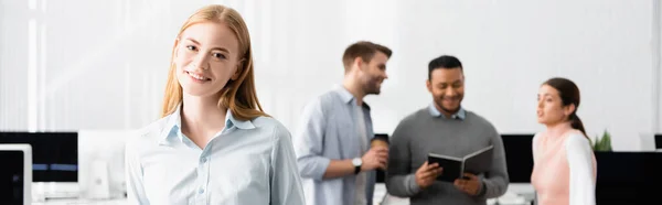 Lächelnde Geschäftsfrau blickt in die Kamera, während multiethnische Kollegen im Hintergrund im Büro arbeiten, Banner — Stockfoto