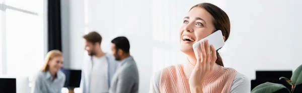 Fröhliche Geschäftsfrau spricht auf Smartphone neben Kollegen auf verschwommenem Hintergrund im Büro, Banner — Stockfoto